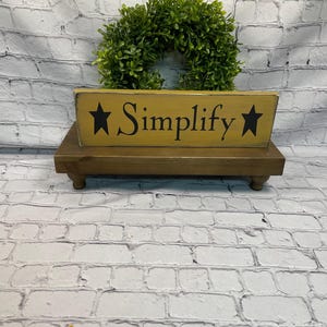 May include: A wooden sign with the word "Simplify" in black lettering, flanked by black stars, sits on a small, brown, wooden stand. A green wreath is behind the sign. The background is a white brick wall.