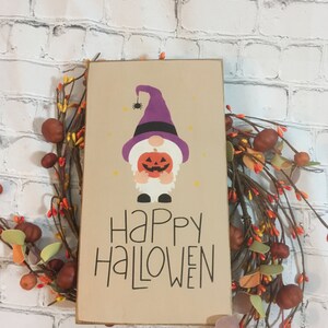 May include: A wooden sign with a brown background featuring a cartoon gnome wearing a purple hat and holding a jack-o-lantern. The sign says "Happy Halloween" in black script.