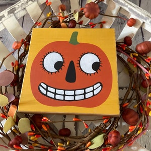 May include: A wooden sign with a painted orange pumpkin with a wide smile and large eyes. The pumpkin is on a yellow background.