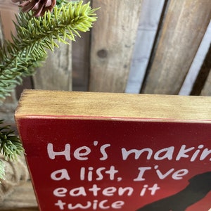 May include: Red wooden sign with white text that reads "He's making a list, I've eaten it twice". A black silhouette of a dog is in the bottom right corner of the sign.