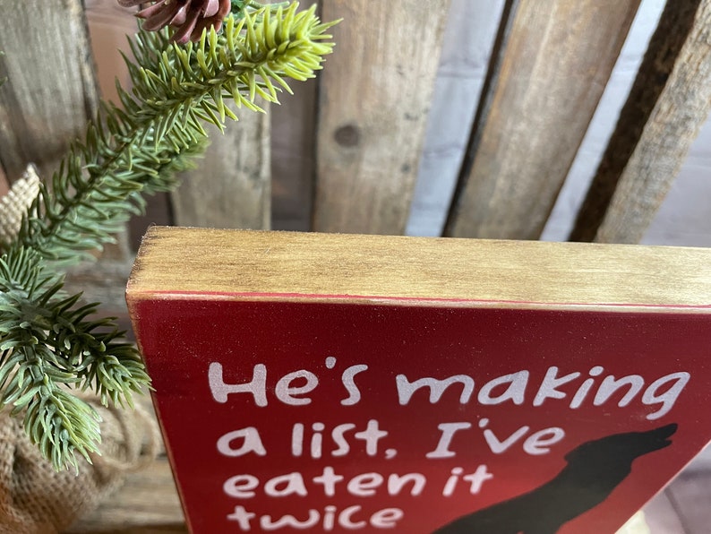 May include: Red wooden sign with white text that reads "He's making a list, I've eaten it twice". A black silhouette of a dog is in the bottom right corner of the sign.