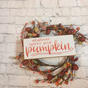 May include: A white wooden sign with the text "whatever spices your pumpkin" in orange lettering. The sign is surrounded by a fall-themed wreath with brown, orange, and yellow leaves and berries.