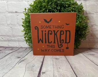 Puede incluir: Un letrero de madera con temática de Halloween. El letrero es naranja y presenta murciélagos negros, una araña y el texto "Something Wicked This Way Comes."