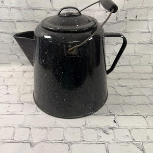 May include: A black enamel coffee pot with a white speckled pattern. The pot features a matching lid, a side handle, and a metal bail handle with a black grip. It has a wide base and a tapered body with a prominent spout on the left side.