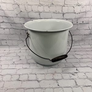 May include: A white enamel bucket with a smooth, glossy finish and a wide, open top. The bucket features a thin, dark metal wire handle that is arched over the top, equipped with a small, dark, cylindrical wooden grip for comfortable handling.