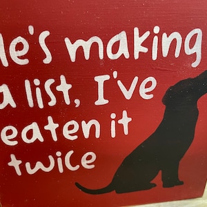 May include: Red wooden sign with white text that reads "He's making a list, I've eaten it twice". A black silhouette of a dog is on the right side of the sign.
