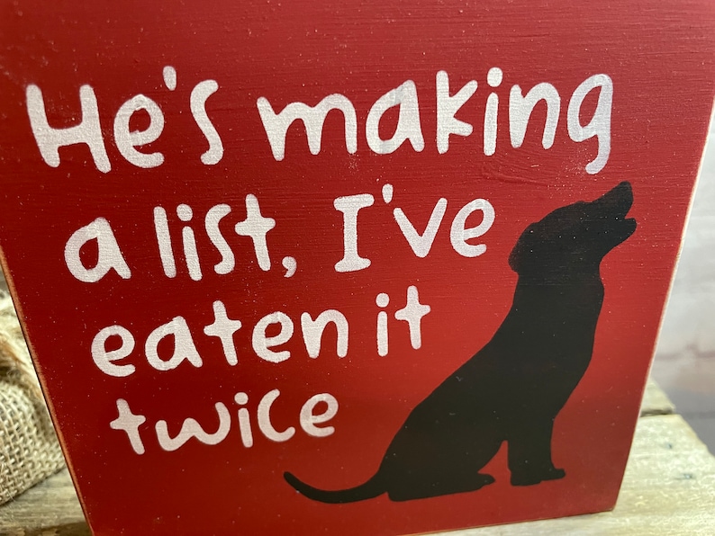 May include: Red wooden sign with white text that reads "He's making a list, I've eaten it twice". A black silhouette of a dog is on the right side of the sign.