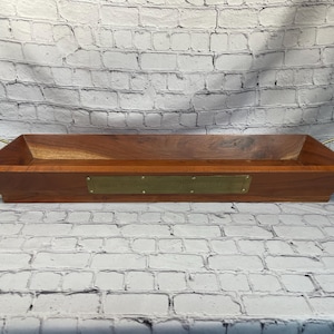 May include: A rectangular wooden serving tray with a rich brown finish. It features brass handles and a brass nameplate. The tray's design is angled at the ends, adding a unique touch to its overall aesthetic.