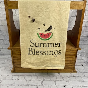 May include: A beige kitchen towel with the words "Summer Blessings" printed in black. The towel features a watermelon slice with a black bird and stars. The towel is displayed on a wooden rack.