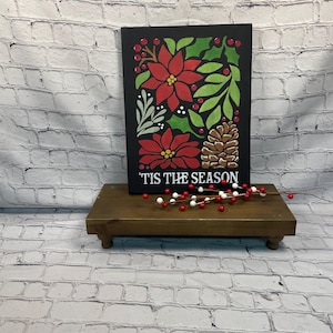 May include: A black rectangular sign with a floral design featuring red poinsettias, green leaves, and a pinecone. The text "'TIS THE SEASON" is written in white. The sign is displayed on a small wooden stand with red and white berries.
