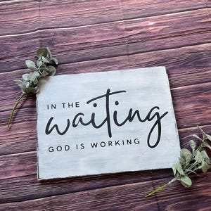 May include: White wooden sign with black text that reads "In the waiting, God is working". The sign is on a woodgrain background.