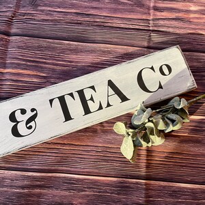 May include: White wooden sign with black text that reads "Cake & Tea Co.". The sign is on a brown wooden background.