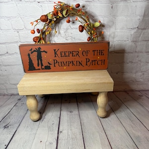 May include: A rectangular orange wooden sign with black text reading Keeper of the Pumpkin Patch and a black silhouette of a scarecrow, resting on a light-colored wooden riser with four turned legs, positioned in front of a decorative autumn wreath.