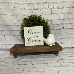 May include: White wooden sign with black calligraphy that reads "Grow in Grace". The sign is sitting on a brown wooden shelf with a white bird figurine to the right.