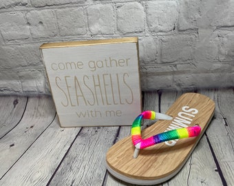 May include: A wooden sign with the text "come gather SEASHELLS with me" next to a flip-flop. The flip-flop has a rainbow-colored strap and the word "SUMMER" printed on the wooden sole.