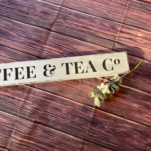 May include: White wooden sign with black lettering that reads "COFFEE & TEA CO" on a brown wood background.