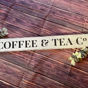 May include: White wooden sign with black lettering that reads "COFFEE & TEA CO". The sign is on a brown wooden background.