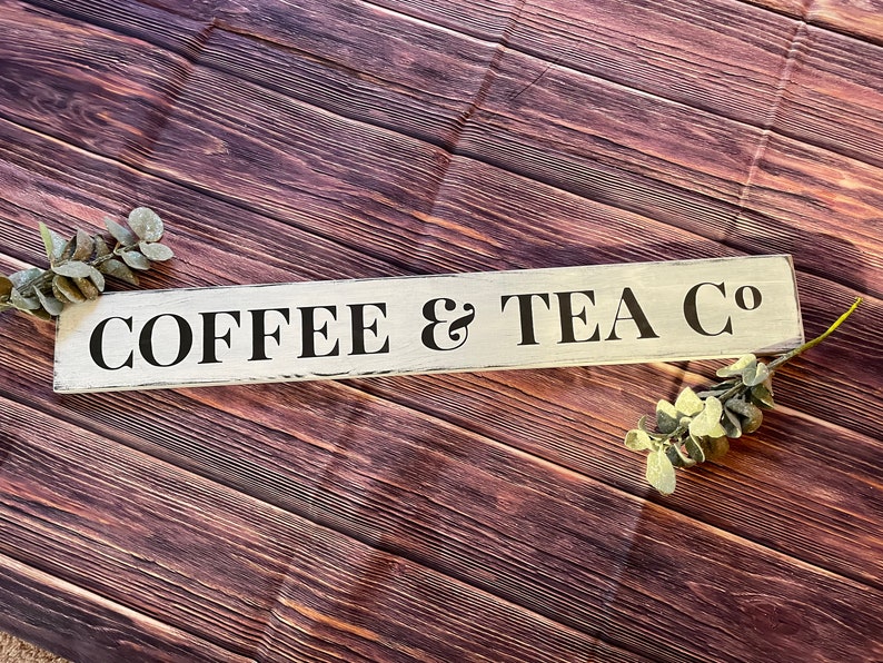 May include: White wooden sign with black lettering that reads "COFFEE & TEA CO". The sign is on a brown wooden background.