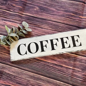 May include: White wooden sign with black lettering that reads "COFFEE &" on a brown wood background with a sprig of greenery.
