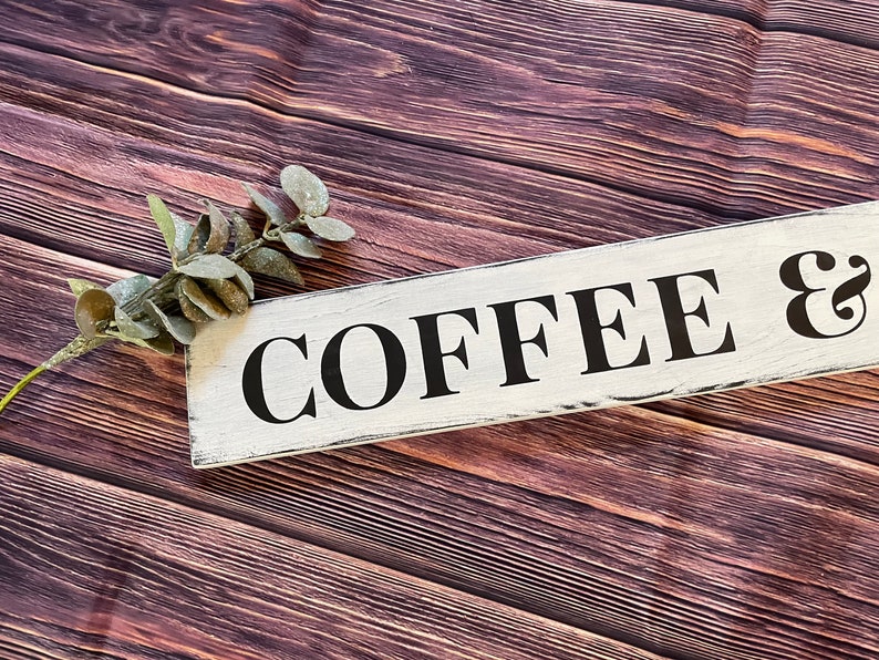 May include: White wooden sign with black lettering that reads "COFFEE &" on a brown wood background with a sprig of greenery.