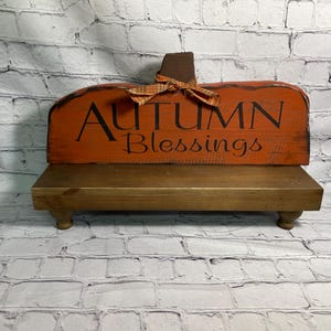 May include: Wooden pumpkin-shaped sign with the words "AUTUMN Blessings" in black lettering. The sign is orange with a brown stem and a plaid bow. It sits on a small, brown wooden stand.