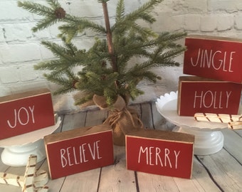 May include: Five wooden blocks with red paint and white lettering. The blocks spell out "Jingle", "Holly", "Joy", "Believe", and "Merry".