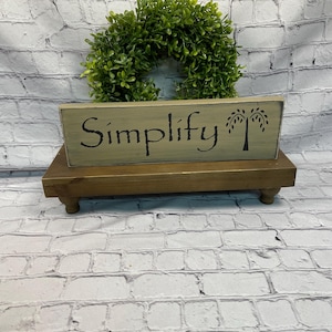 May include: A wooden sign with the word "Simplify" in black script, accompanied by a tree silhouette. The sign is placed on a small, brown, wooden riser with rounded feet. A green wreath sits behind the sign against a white brick wall.