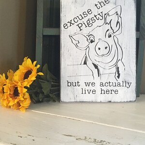 May include: White wooden sign with black text and a cartoon pig. The text reads "Excuse the Pigsty but we actually live here."