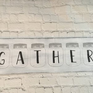 May include: White wooden sign with the word "GATHER" spelled out in black letters. Each letter is inside a gray silhouette of a mason jar.