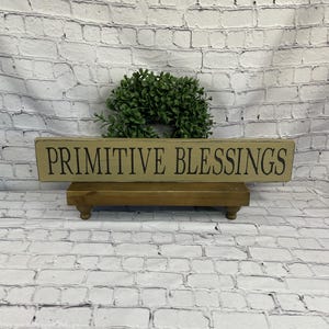 May include: A rectangular wooden sign with the words "PRIMITIVE BLESSINGS" in black lettering. The sign is placed on a small, brown wooden stand. A green wreath is behind the sign, and the background is a white brick wall.