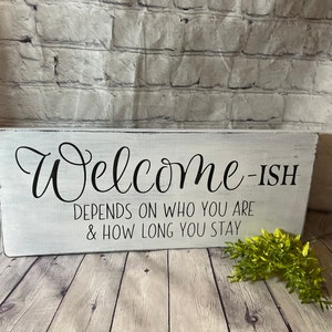 May include: White wooden sign with black text that reads "Welcome-ish Depends on who you are & how long you stay".