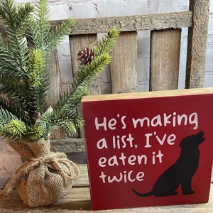 May include: Red wooden block sign with white text that reads "He's making a list, I've eaten it twice". A black silhouette of a dog is on the right side of the sign.