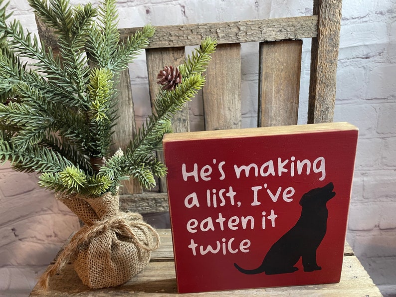 May include: Red wooden block sign with white text that reads "He's making a list, I've eaten it twice". A black silhouette of a dog is on the right side of the sign.