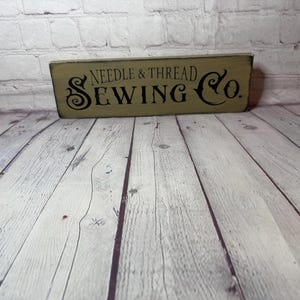 May include: A rectangular wooden sign with the text "NEEDLE & THREAD SEWING Co." in black lettering. The sign has a distressed, aged appearance with a light green background. The sign is placed on a wooden surface.