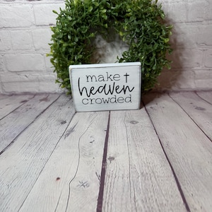 May include: A white wooden sign with the text "make heaven crowded" in black script, placed in front of a green wreath. The sign is on a light-colored wooden surface.