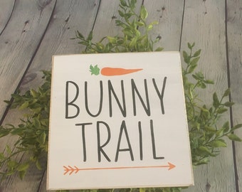 May include: White wooden sign with the text "Bunny Trail" in black lettering. A small orange carrot with a green stem is above the text. An orange arrow points to the right.