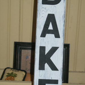 May include: White wooden sign with black painted letters spelling "BAKE".
