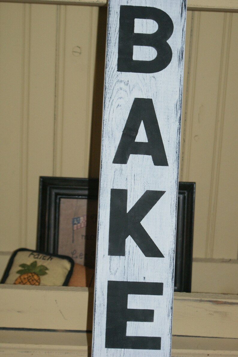 May include: White wooden sign with black painted letters spelling "BAKE".