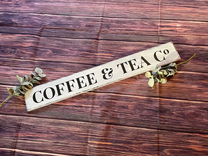 May include: White wooden sign with black lettering that reads "COFFEE & TEA CO" on a brown wood background.
