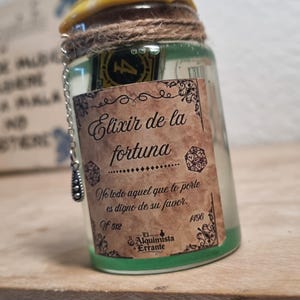 Puede incluir: Una pequeña botella de vidrio con tapa amarilla y etiqueta decorativa. La etiqueta dice "Elixir de la fortuna" y contiene texto en español. La botella está sellada con hilo y tiene una pequeña cadena adjunta.