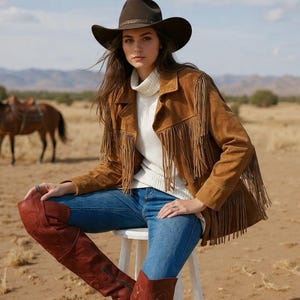 May include: A brown suede jacket with long fringe detailing on the chest and sleeves, worn over a white ribbed turtleneck sweater, blue denim jeans, and tall brown leather boots, paired with a dark brown felt cowboy hat.