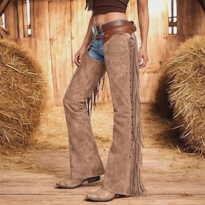May include: Tan suede leather chaps featuring long fringe along the outer legs, a brown leather belt with a silver buckle, and metal studs. The chaps are worn over blue denim jeans, paired with a brown crop top and tan leather pointed-toe boots.
