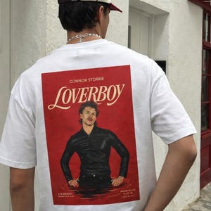 May include: White t-shirt with a red graphic on the back. The graphic features the word "Loverboy" and a portrait of a man. The shirt also has the name "Connor Storrie" printed above the word "Loverboy". The person is wearing a baseball cap.