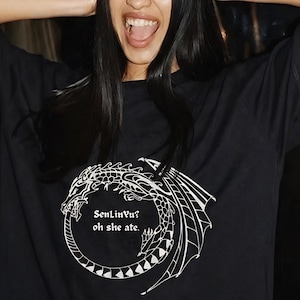 Op de afbeelding: Zwart T-shirt met een witte drakenafbeelding en de tekst "SenLinVu? oh she ate." Het drakenontwerp heeft gedetailleerde vleugels en een cirkelvormig lichaam. Een casual kledingstuk.