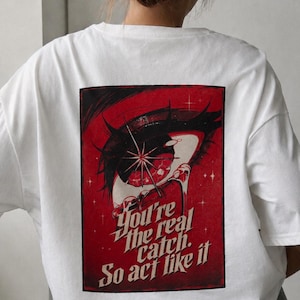 Könnte beinhalten: Ein weißes T-Shirt mit kurzen Ärmeln und einer rechteckigen Grafik auf der Rückseite. Das Design zeigt ein stilisiertes rotes und schwarzes Auge mit einem Sternenlicht-Effekt und dem Text: You're the real catch. So act like it, in fetter, kursiver Schrift.