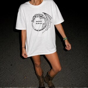 Op de afbeelding: Wit T-shirt met een zwart drakenontwerp en de tekst "best life oh she ate". Het shirt wordt gedragen met bruine cowboylaarzen. De achtergrond is donker.