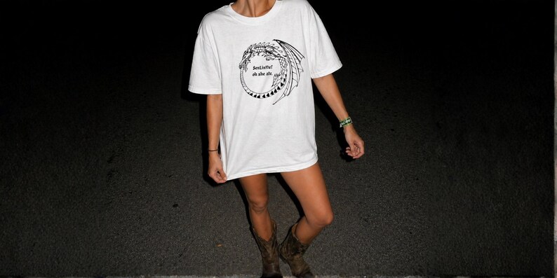 Op de afbeelding: Wit T-shirt met een zwart drakenontwerp en de tekst "best life oh she ate". Het shirt wordt gedragen met bruine cowboylaarzen. De achtergrond is donker.