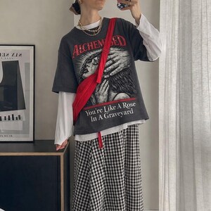 May include: A person wearing a dark gray graphic t-shirt with the text "ALCHEMIZED" and "You're Like A Rose In A Graveyard". The outfit includes a white long-sleeve shirt, a red fanny pack, and a black and white checkered skirt.