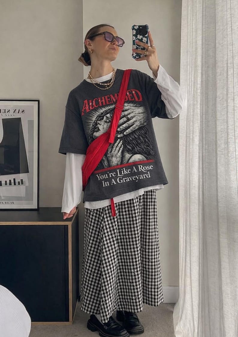May include: A person wearing a dark gray graphic t-shirt with the text "ALCHEMIZED" and "You're Like A Rose In A Graveyard". The outfit includes a white long-sleeve shirt, a red fanny pack, and a black and white checkered skirt.