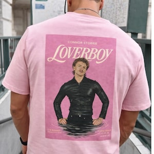 May include: Light pink t-shirt featuring a graphic of a man in a black leather jacket with the word "Loverboy" above the image. The text "CONNOR STORRIE" is above the word "Loverboy". The shirt is light pink.